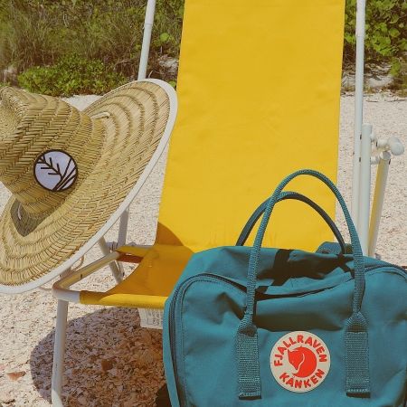Tropii Co. sunhat on a yellow beach chair at the beach with foliage in the background and a teal blue Fjallraven Kanken backpack.