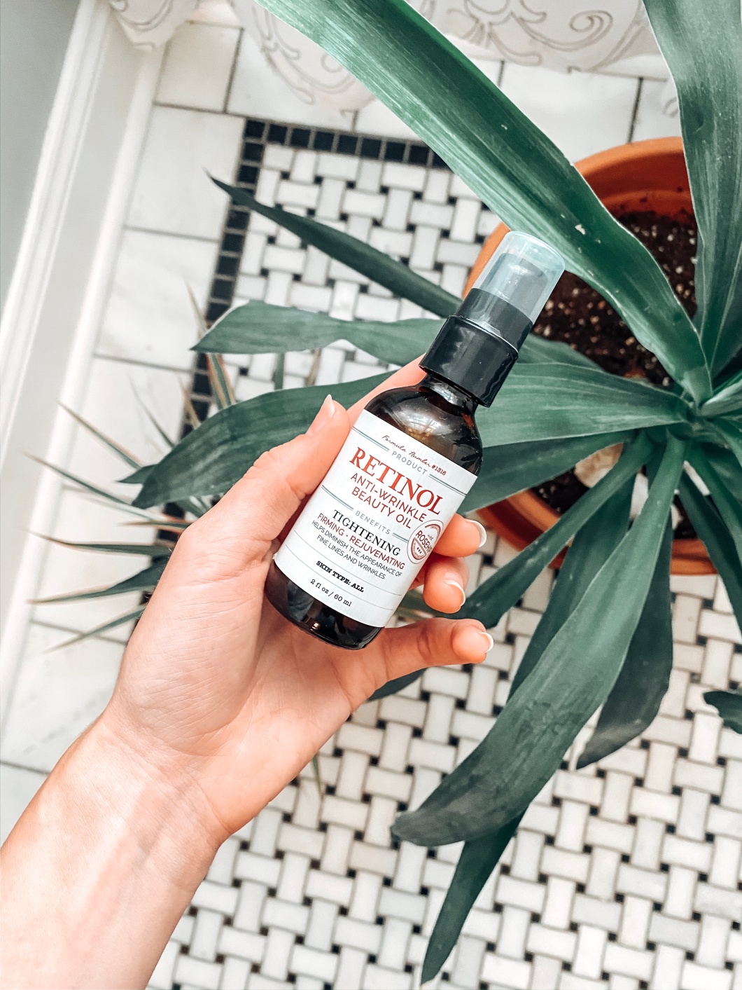 Image of Candace's hand holding a bottle of Retinol Oil over plants and black and white marble tile.